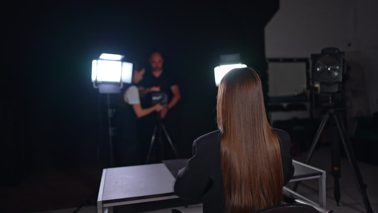 Unrecognized long-haired woman sits at desk in the light of soffits. Cameraman and female assistant work backstage.