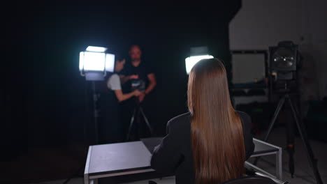 unrecognized long-haired woman sits at desk in the light of soffits. cameraman and female assistant work backstage.