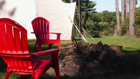 two chairs stand near a bonfire in the forest in the daytime, while a tent is established in the background