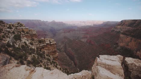 Grand-Canyon-cliff-edge-gimbal-push-forwards-reveals-valley-below,-with-blue-skies,-cloud-cover,-soft-midday-sun,-red-rocks,-and-shadows-on-the-canyon-walls---Arizona-USA