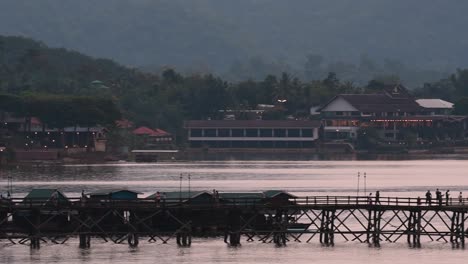 Mon-Bridge-and-a-longboat-followed-speeding-behind-the-structure-while-people-are-walking-on-the-bridge,-silhouetting-as-it-was-getting-dark,-in-slow-motion