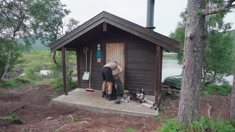 shirtless man walk towards the cabin to unpack with dog lying on the floor