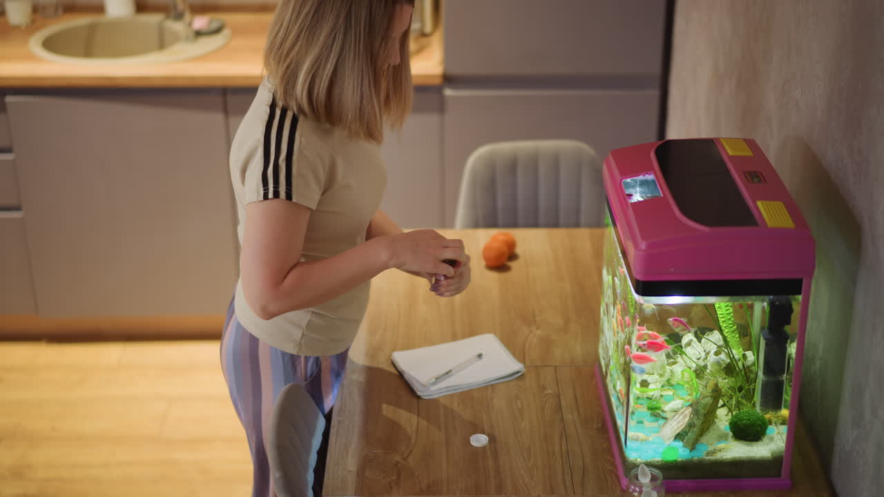 Colorful tropical fish swim inside illuminated pink aquarium on wooden table beside notebook with pen, orange fruit, small jar, and person partially visible, creating warm indoor workspace