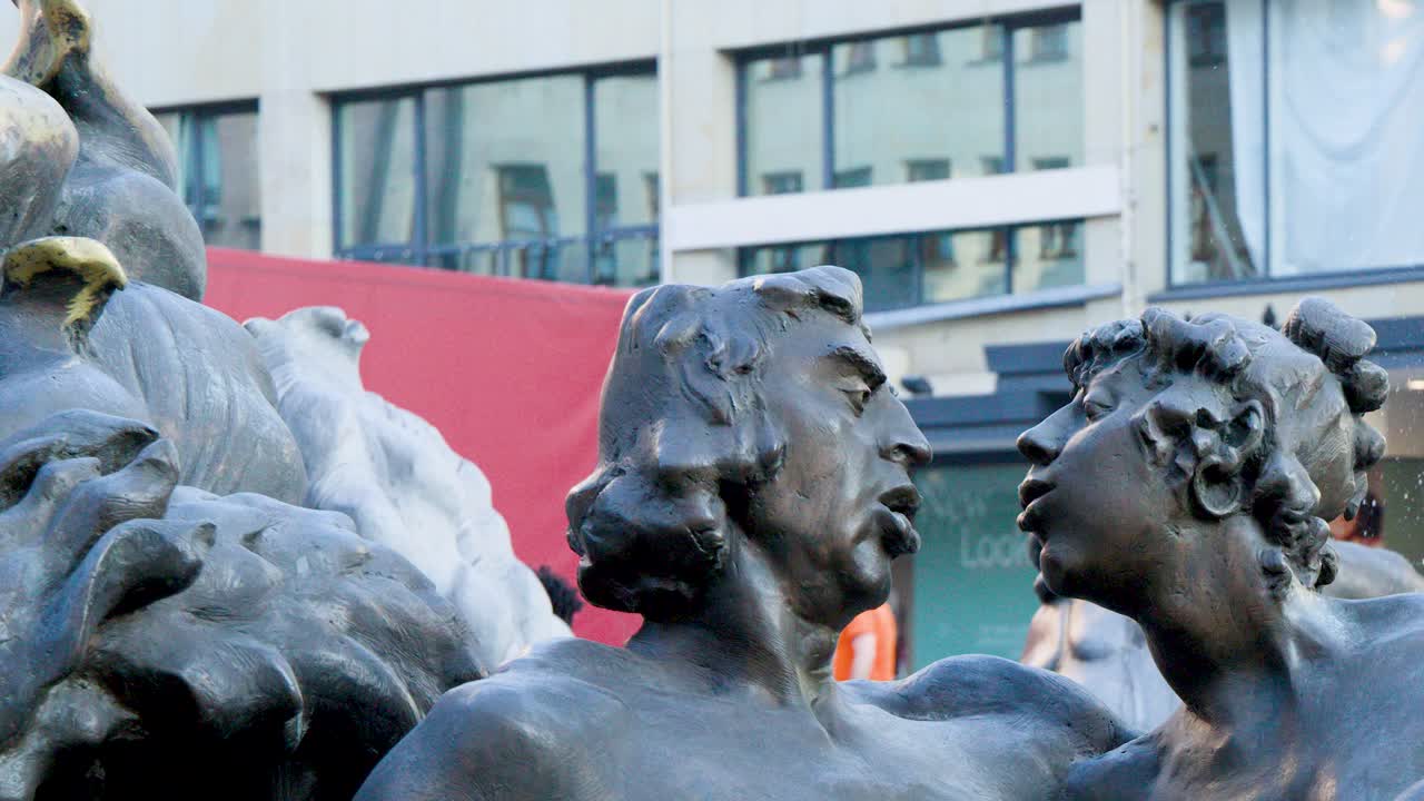 Bronze statues in a public fountain, water splashing, modern urban background, daylight, static shot