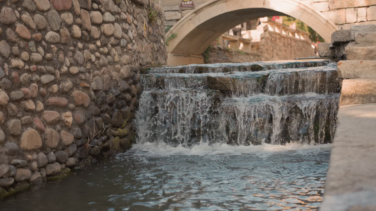 Cascata su un ponte di pietra che scorre sui gradini, la morbida luce serale riscalda i muri in muratura invecchiati, leggere increspature e una caduta spumosa in una conca poco profonda, atmosfera urbana tranquilla con sottili riflessi sull'acqua.