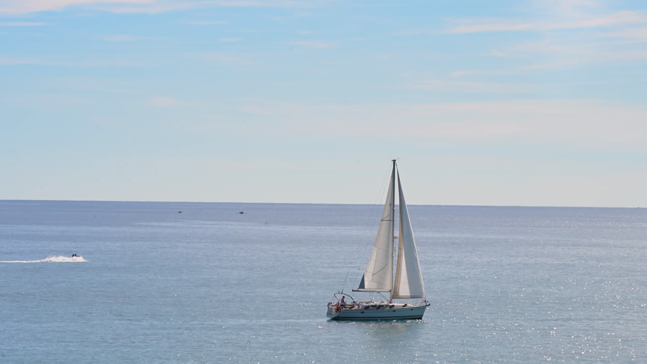 A sailboat with full white sails glides across the calm Mediterranean Sea near Alicante, Spain. In the background, a person on a jet ski enjoys the water at a distance