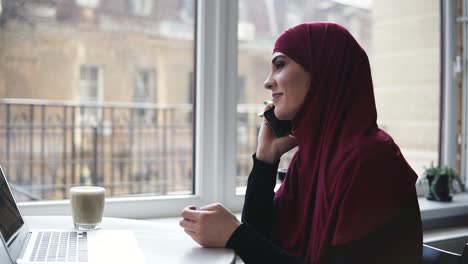 young attractive girl of unrecognizable nationality with hijab on her head is talking to someone on the phone while she has laptop and glass of cappuccino lying in front of her