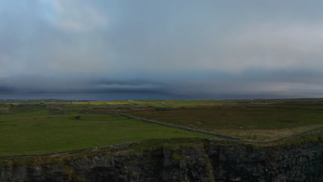 Tiro-Ascendente-Del-Acantilado-Rocoso.-Revelando-Verdes-Prados-Y-Pastos-Y-El-Mar-En-La-Distancia.-Cielo-Nublado.-Acantilados-De-Moher,-Irlanda