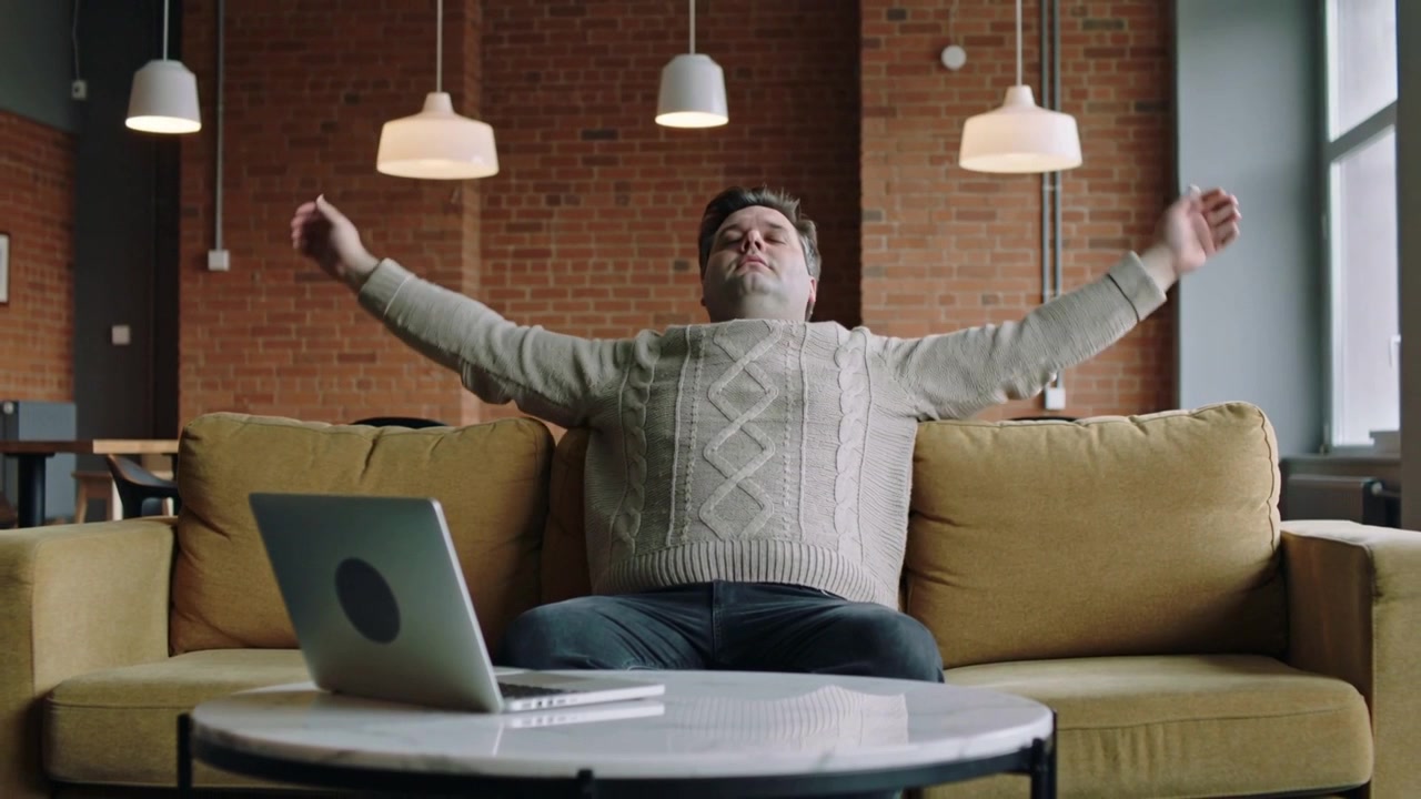 Stretching man in turtleneck taking short break from laptop on sofa at home then leaning back
