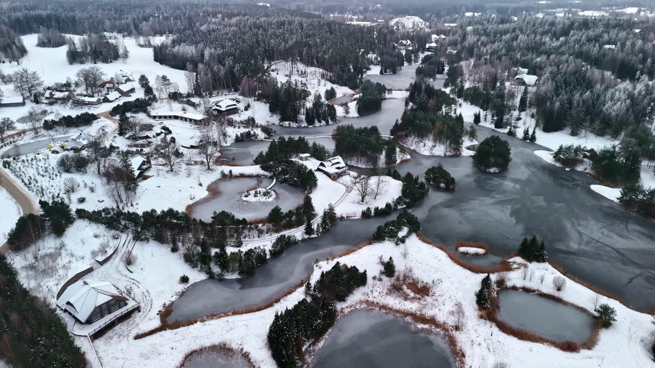 Smooth aerial pan across snow-blanketed forest, revealing cozy cabins, winding paths, and a chain of frozen ponds in winter