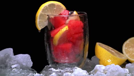 glass filled with watermelon cubes and lemon, surrounded by sliding ice on black screen, water drop falling from lemon