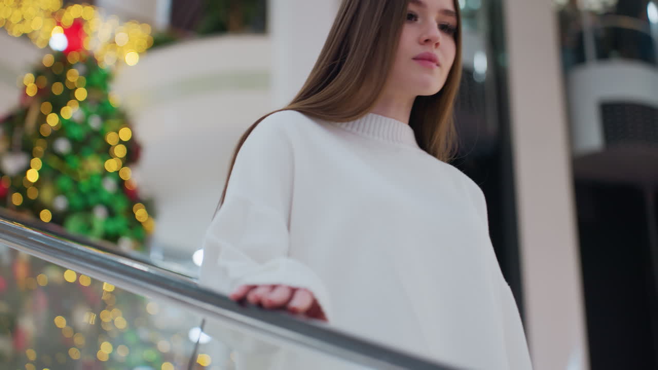 Lady calmly descending escalator, hand on rail, in beautifully decorated shopping mall with large illuminated Christmas tree in background