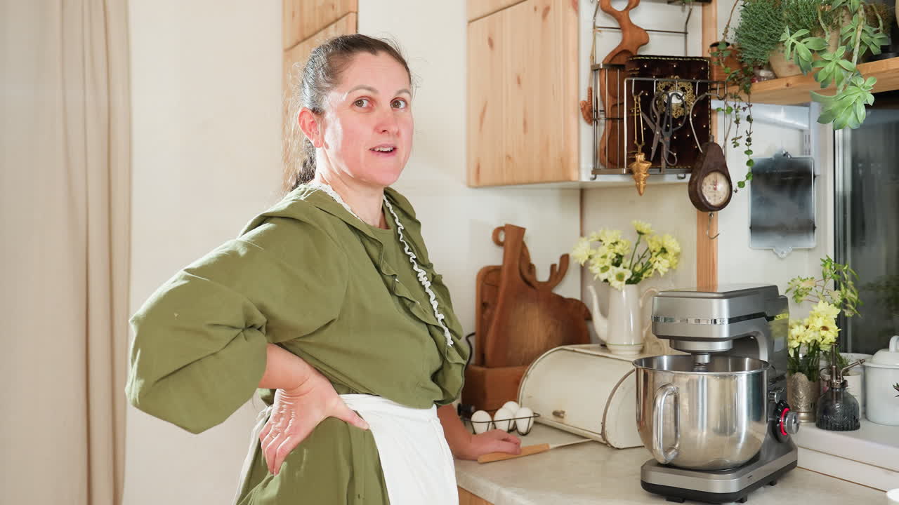 Middle aged woman in green dress and white apron stands in cozy kitchen beside electric stand mixer, smiling warmly at camera with hand on waist while waiting for mixing process to complete