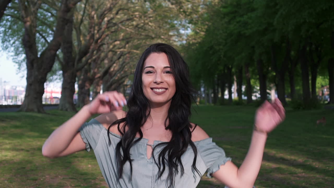 atractiva y juguetona mujer latina con cabello negro ondulado caminando bajo los árboles en un parque en londres, mirando a la cámara, girando feliz con una hermosa sonrisa