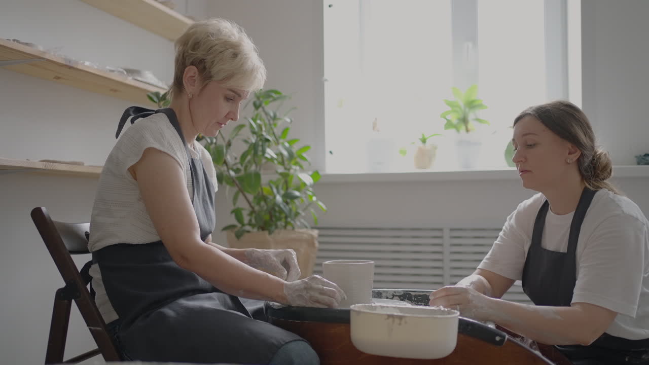 Woman master transfers knowledge to an elderly woman working on a potter's wheel and making a mug of ceramics in her workshop in slow motion