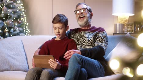 portrait of happy smiling old grandpa with teen grandchild sitting in decorated room typing and browsing on tablet while chatting near glowing new year tree