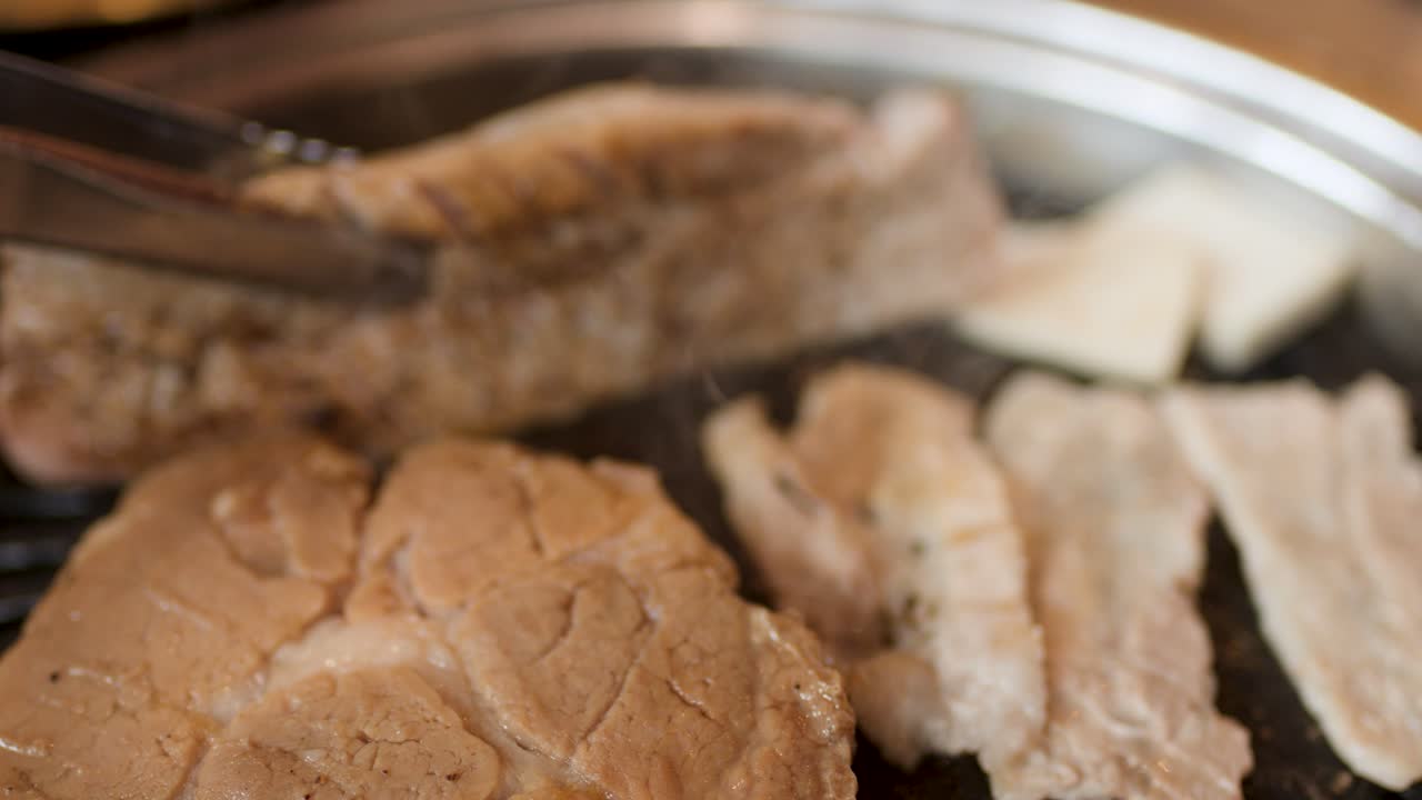 Tongs lift grilled pork from hot Korean barbecue grill, warm lighting, shallow depth of field
