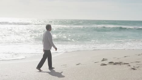 Side-view-of-hispanic-senior-man-walking-on-beach-at-sunset