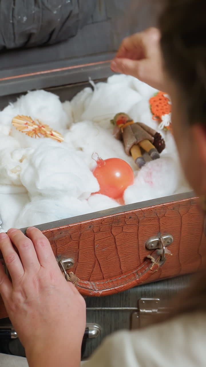 흰색 옷을 입은 여성이 여행 가방을 열어 장신구를 찾고, 오렌지색 유리 구슬이 면 둥지에서 살짝 보입니다. 나무 조각상은 부드러운 패딩 옆에 놓여 있고, 손은 조심스러운 리듬으로 장식품을 정리하며 트리에 대한 즐거운 기대를 표현합니다.