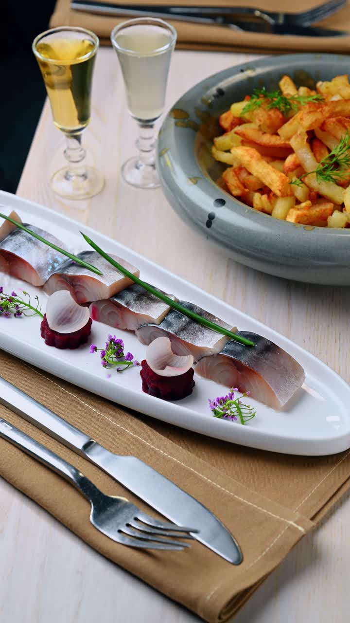 Tavola completamente apparecchiata con antipasto di aringhe e contorno di patate. Piatto lungo con fette di pesce, ciotola di patatine fritte e bicchierini di liquore su un tavolo di legno chiaro.