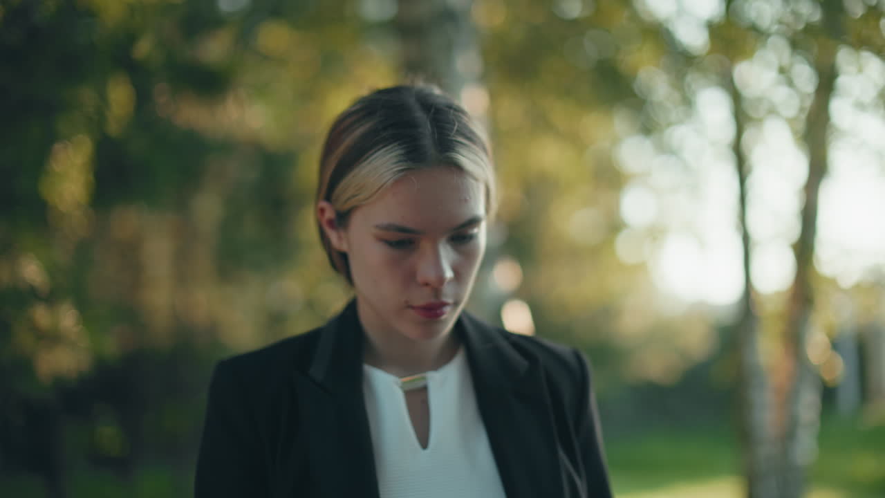 Professional woman enjoys coffee in tranquil park setting with trees behind her as warm sunlight filters through leaves, casting soft natural light across her focused face during outdoor break