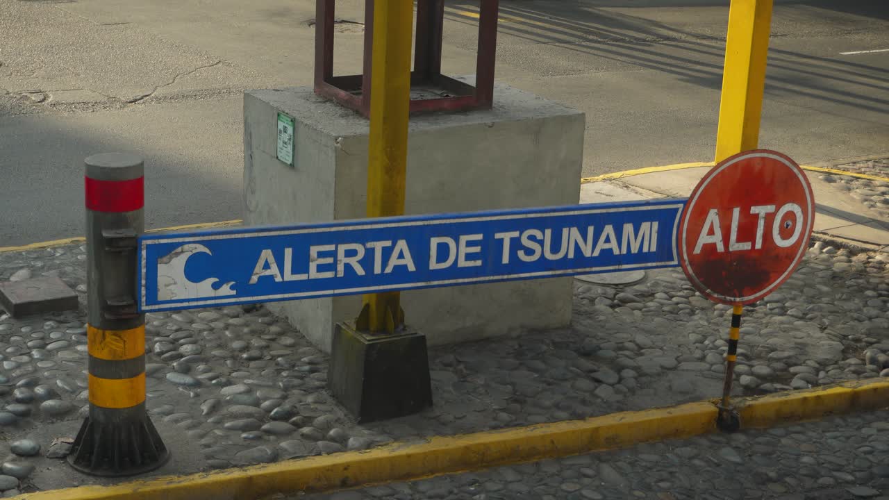 Una señal de advertencia de tsunami junto a una señal de stop. Ambas escritas en español. Coches pasando por detrás. Ubicado en el distrito de Miraflores de Lima, Perú.