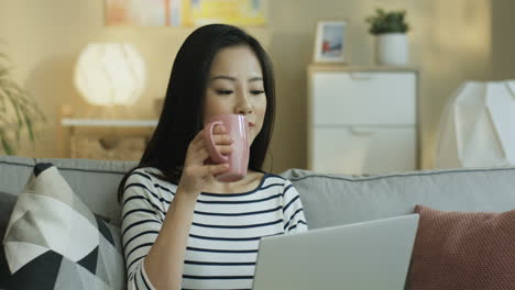 줄무늬 셔츠를 입은 젊은 여성이 커피를 마시고 거실 소파에 앉아 노트북에서 무언가를 보고 있습니다.