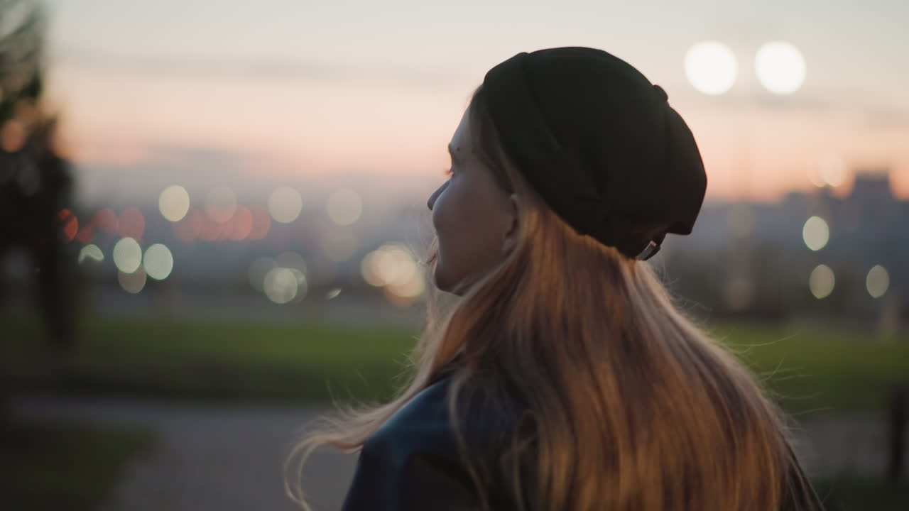 back view young woman overlooking city wearing beanie and jacket headphones around neck long hair cascading with distant bokeh lights at dusk, contemplative silent mood, urban overlook, cinematic