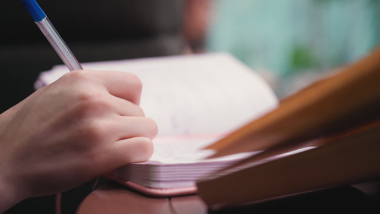 Close up view of woman hand holding blue pen while writing in open notebook, background blurred with soft tones and partially visible book on side creating calm and focused workspace vibe