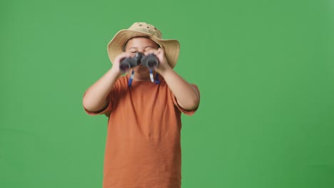 asian tourist boy with a hat wipes his sweaty face after looking through the binoculars. boy researcher examines something on the green screen background, travel tourism adventure concept