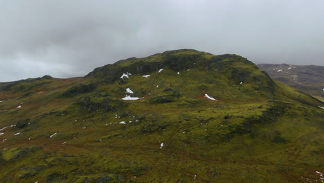Zoom-En-La-Vista-De-Drones-De-Una-Formación-Rocosa-En-La-Ladera-De-Una-Montaña-Que-Se-Asemeja-A-Un-Cráter-Rodeado-De-Cielos-Nublados-Y-Montañas-En-La-Distancia-En-Islandia