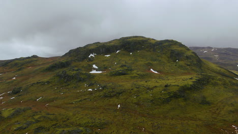 Zooming-in-drone-view-of-a-rock-formation-on-the-side-of-a-mountain-resembling-a-crater-surrounded-by-overcast-skies-and-mountains-in-the-distance-in-Iceland