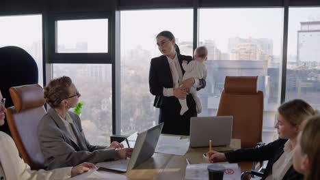 una confiada chica morena con gafas redondas en un traje de negocios negro sostiene a un niño pequeño en sus manos y tiene una reunión de mujer de negocios en una oficina moderna en la mesa