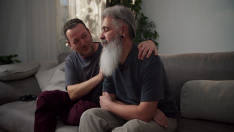 a sad man with gray hair and a lush beard with a black earring in his ear and in a gray t-shirt is sad and his middle-aged brunette boyfriend supports him sitting on the sofa in a modern apartment in the evening
