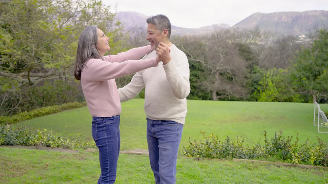 Senior couple clasping hands, stepping on brick patio, twirling on lawn and hugging while laughing