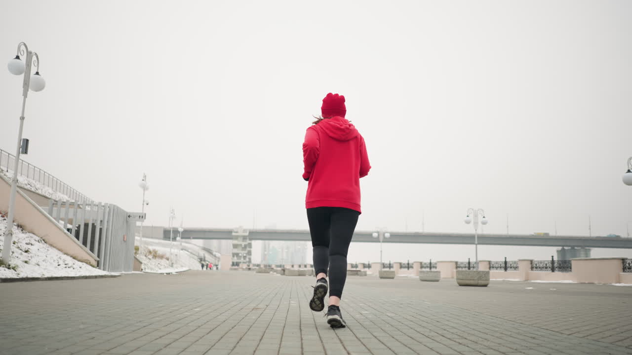 vista trasera de un atleta corriendo al aire libre en el pavimento entrelazado en invierno con suelo cubierto de nieve, elementos urbanos modernos, puente con coche en movimiento, postes de luz y bancos