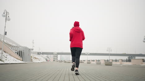 back view of athlete jogging outdoors on interlocked pavement in winter with snow-covered ground, modern urban elements, bridge with moving car, lamp posts, and benches