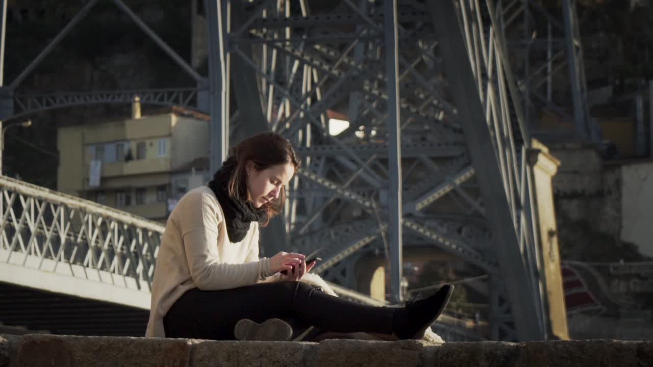 Splendida signora che usa lo smartphone mentre è seduta vicino alla struttura del ponte in acciaio a Porto, Portogallo - ripresa in super slow motion