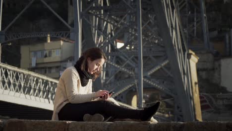 gorgeous lady using smartphone while sitting by the steel bridge structure in porto, portugal - full slowmo shot