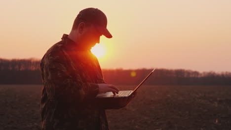 Ein-Bauer-Mittleren-Alters-Benutzt-Einen-Laptop-Bei-Sonnenuntergang-Auf-Einem-Feld-Und-Plant-Die-Aussaat