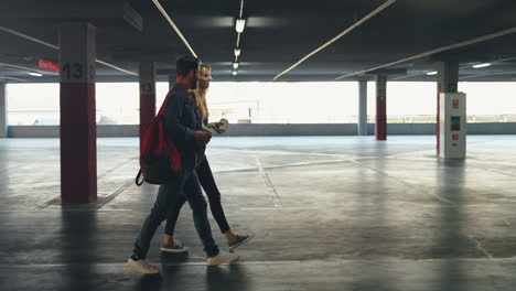 Vista-Lateral-De-Un-Joven-Hombre-Y-Una-Mujer-Caucásicos-Y-Elegantes-Caminando-En-Un-Estacionamiento-Vacío-Y-Hablando-Mientras-Beben-Café