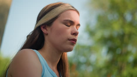 young woman outdoors, gently touching her hair while sunlight softly illuminates her face, wearing cyan top, with hair pinned back using black pin, under blue sky, surrounded by blurred greenery