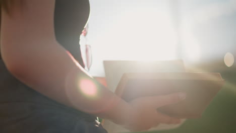 la luz del sol brilla de la vegetación borrosa en el fondo, reflejándose en un libro sostenido por una mujer sentada al aire libre, ella revisa suavemente las páginas mientras lleva gafas en su camiseta negra
