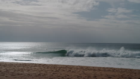 Sand-beach-in-Nazare,-Portugal,-waves-and-few-surfers,-static-afternoon-view-of-Atlantic-ocean