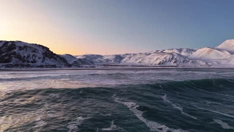 aerial drone gliding and flying over dark cold ocean sea water with mountains in the background during sunset or sunrise with orange sky in norway during winter. drone reaches the beach dark shoreline