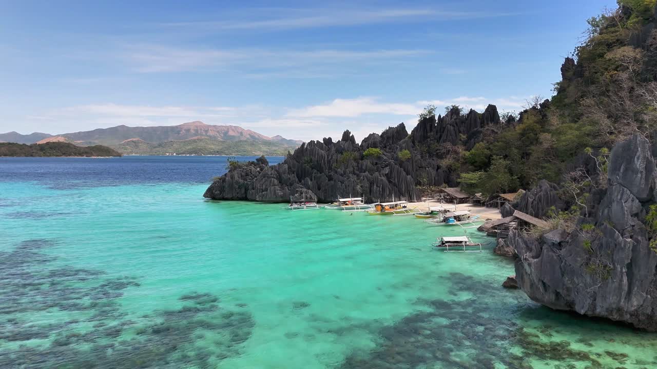 Turquoise waters meet white sand and dramatic limestone formations at Smith Point Beach in Coron, Philippines, with traditional boats anchored near the shore and lush greenery surrounding the beach