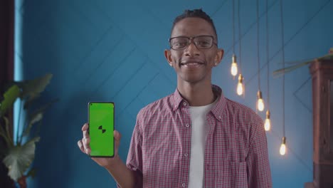 black man with glasses in the room holds phone with an app in hand and smiles, green screen. black man smiling shows a phone screen a mockup