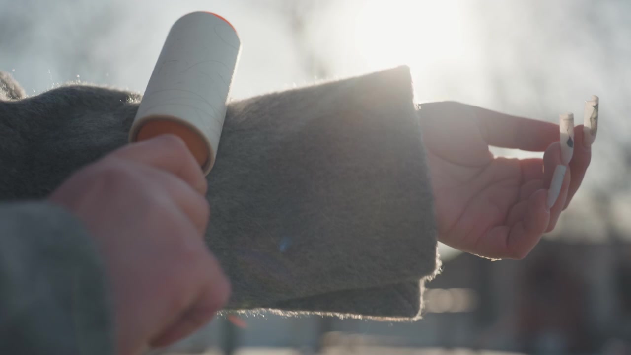 Mani che passano un rullo levapelucchi su una manica, cortile invernale in controluce con luce solare soffusa, primo piano su fibre di tessuto e unghie dipinte, movimento delicato rimuove pelucchi e detriti, routine mattutina accogliente e tattile.