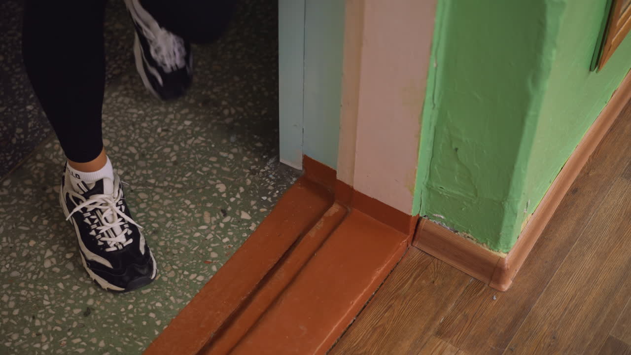 Close leg view of lady in black leggings wearing canvas sneakers stepping out through doorway, casual exit captured on threshold with details of green wall and polished wooden floor