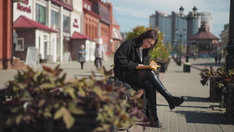 woman seated outdoors reading a book, flipping through pages with her bag on the bench, as people walk by in the blurred background